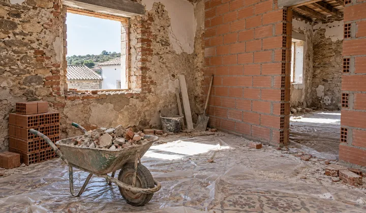 Interior de una casa antigua en rehabilitación: carretilla con escombro, ladrillos apilados, tabique nuevo de ladrillo hueco y suelo hidráulico protegido con plástico